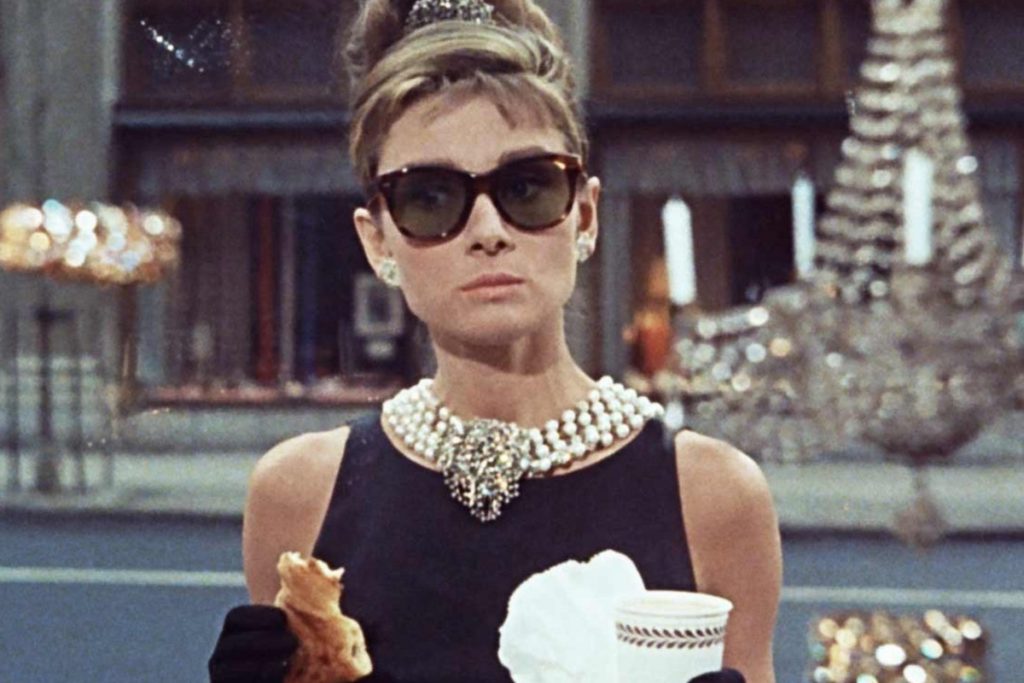 The opening scene: Holly holding a coffee paper cup and a patisserie. She's wearing the iconic black dress and with a triple strand necklace, earrings and a head piece.
